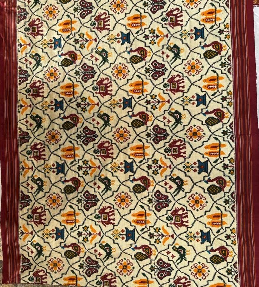 Close-up of cream Patola Ikat fabric featuring a geometric grid with elephant, parrot, and floral motifs, bordered by striped maroon edges.