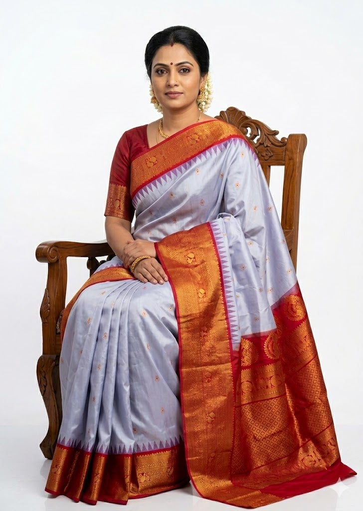 A beautiful traditional Indian woman sitting on a wooden chair against a solid white background, elegantly draped in an authentic pure Gadwal silk saree. The saree features a light blue body with gold zari buttas and a striking contrast red border with intricate gold zari temple motifs. She is wearing a matching red silk blouse and traditional gold jewelry.