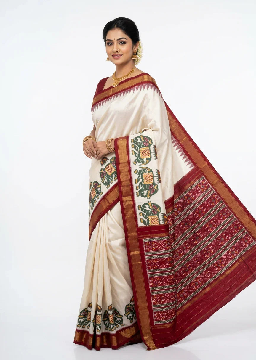 A beautiful Indian woman standing gracefully against a minimal white background, modeling an elegant ivory Pochampally Ikkat silk saree. The saree features a striking deep red and gold zari border with intricate green and yellow elephant motifs. The drape showcases a grand, contrasting red pallu with classic geometric Ikkat weave patterns. She is styled with traditional gold jewelry and a floral hair bun.