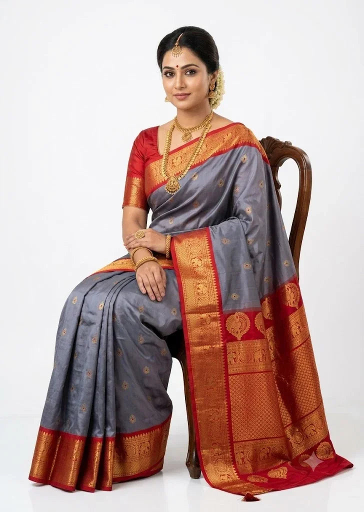 Traditional Indian woman seated on a chair wearing a slate gray Gadwal silk saree with a red zari pallu and red blouse against a plain white background.