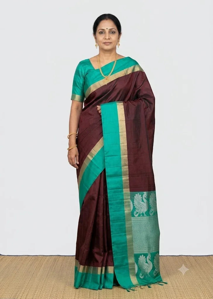 A beautiful Indian woman wearing a traditional deep maroon Kanchi Soft Silk Saree with a high-contrast teal green border featuring complex gold zari and silver peacock motifs.