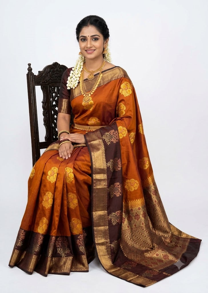 Indian woman wearing a mustard rust semi Dharmavaram silk saree with deep wine zari border, seated on an antique wooden chair against a plain white studio background