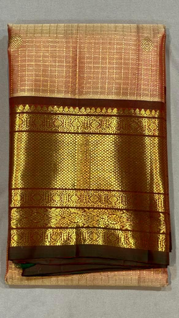 A woman sitting on a wooden chair wearing a traditional Kanchipuram pure silk saree with a peach checkered body and a heavy dark maroon zari border.