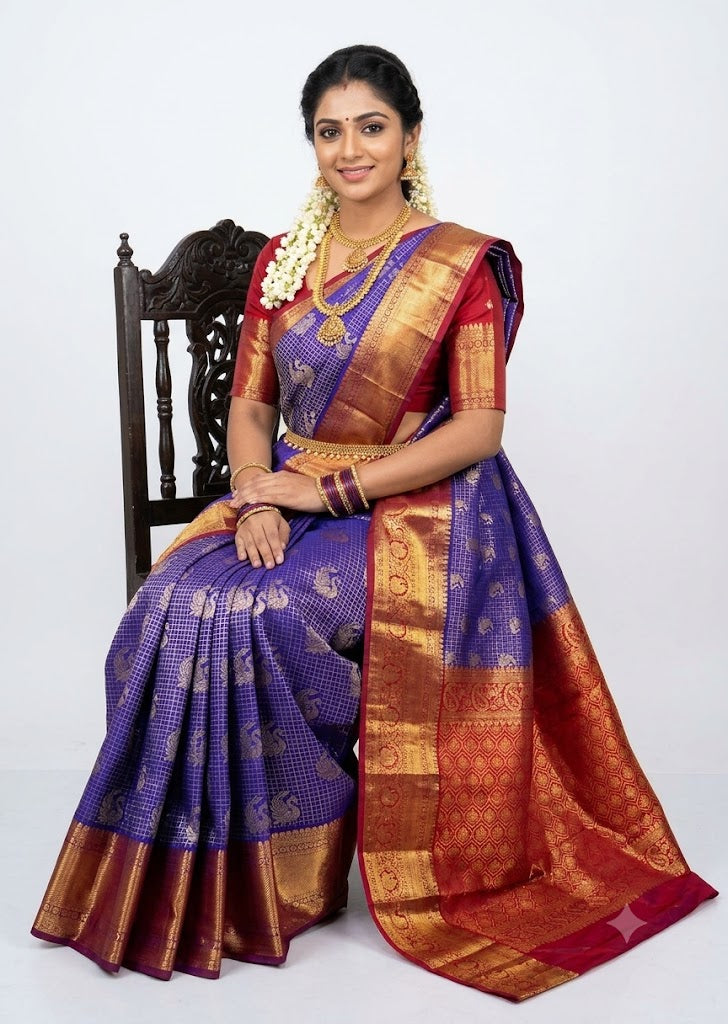Indian woman wearing a royal blue semi Dharmavaram silk saree with red zari border, sitting gracefully on an antique wooden chair against a plain white background.