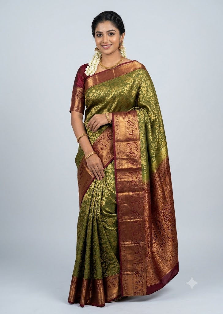 A beautiful traditional Indian woman smiling and posing in an olive green and maroon Dharmavaram silk saree with elaborate gold zari work, featuring a matching maroon blouse, traditional gold jewelry, and jasmine flowers in her hair against a light gray backdrop.