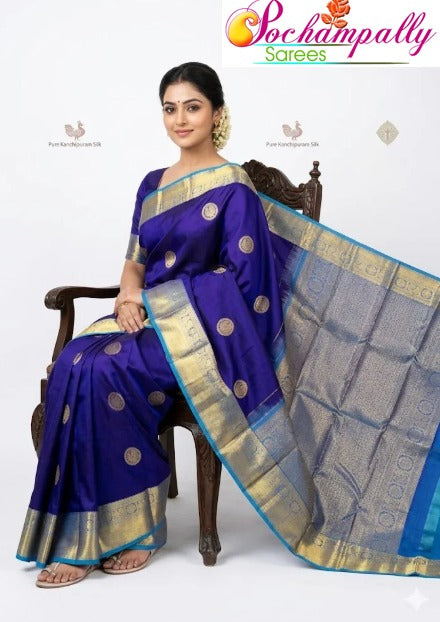 A woman seated on a wooden chair modeling a royal blue Pure Kanchipuram silk saree featuring gold zari buttas across the body, a traditional golden zari border with light blue piping, and an elaborately woven pallu, from Pochampally Sarees.