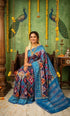 A smiling woman sitting on a traditional wooden swing wearing a vibrant blue Ikkat Patola silk saree featuring intricate purple and orange geometric and animal motifs.
