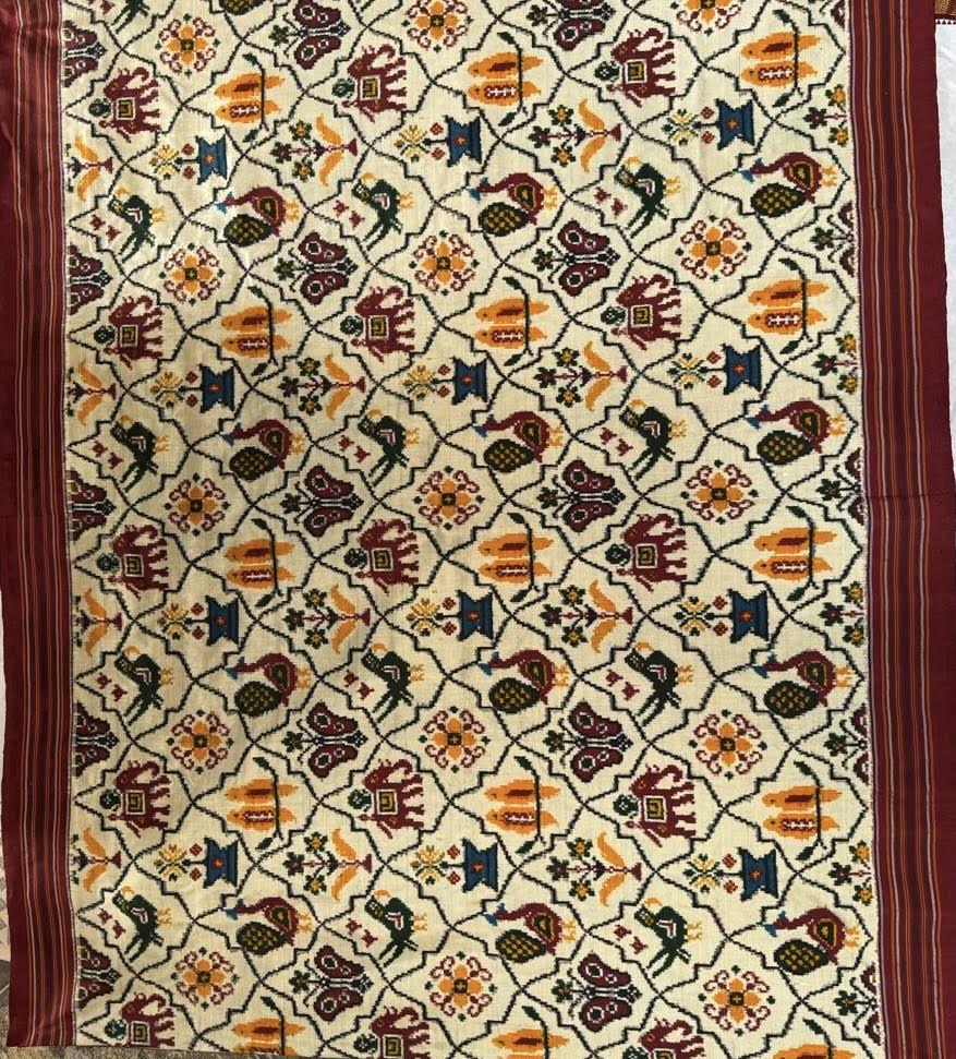 Close-up of cream Patola Ikat fabric featuring a geometric grid with elephant, parrot, and floral motifs, bordered by striped maroon edges.