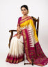 A beautiful traditional Indian woman seated on a carved wooden chair, modeling an off-white and yellow Pochampally Ikkat silk saree with a magenta zari border and intricately woven geometric pallu against a clean white background.
