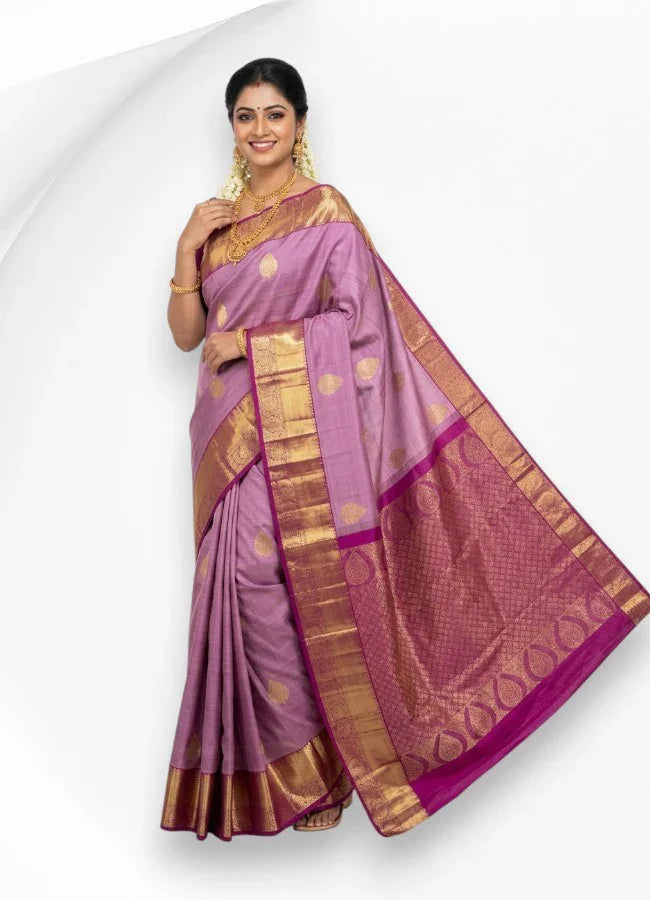 A smiling South Indian woman wearing a traditional pastel lavender kanchi pattu saree with gold motifs and a contrasting magenta gold zari border, styled with traditional temple jewelry and jasmine flowers.