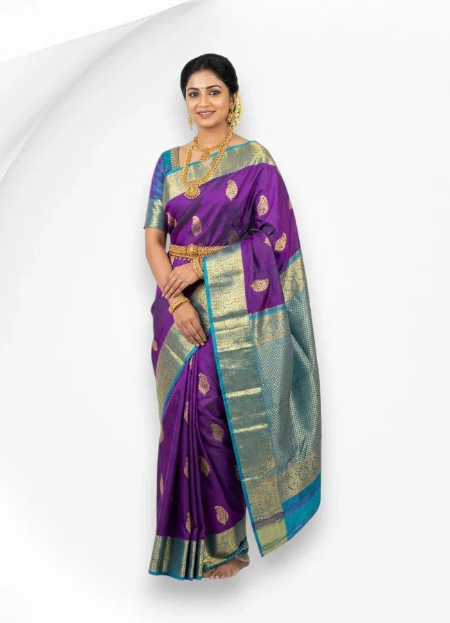 A woman smiling while wearing a rich purple pure Kanchipuram pattu silk saree featuring gold paisley motifs and a contrasting teal blue zari border, styled with traditional gold jewelry.