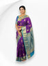 A woman smiling while wearing a rich purple pure Kanchipuram pattu silk saree featuring gold paisley motifs and a contrasting teal blue zari border, styled with traditional gold jewelry.