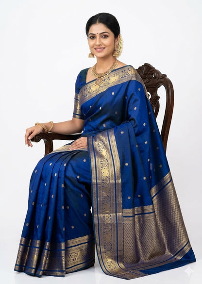 A woman wearing a royal blue Kanchipuram Pattu saree featuring a wide gold zari border and small gold buttas. She is seated in a carved wooden chair and accessorized with traditional gold temple jewelry and a floral hair arrangement.