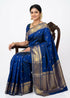 A woman wearing a royal blue Kanchipuram Pattu saree featuring a wide gold zari border and small gold buttas. She is seated in a carved wooden chair and accessorized with traditional gold temple jewelry and a floral hair arrangement.