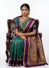 Indian woman seated on a carved wooden chair wearing a teal green pure Kanchipuram silk saree with a contrast purple border and intricate gold zari work, styled with heavy gold temple jewelry against a white background.