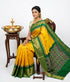 A beautiful traditional Indian woman seated on a wooden chair, modeling a bright yellow Pochampally Ikkat silk saree with an emerald green zari border and intricate geometric diamond pallu. She is wearing traditional gold jewelry and jasmine flowers in her hair, positioned next to a brass diya and floral bowl against a clean white background.