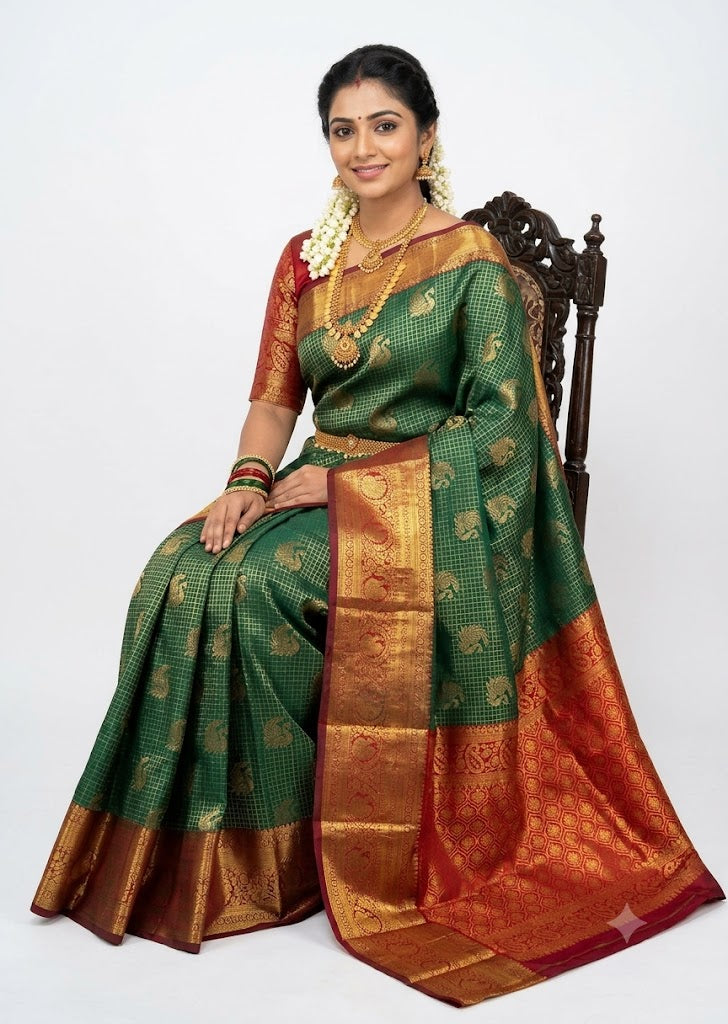Indian woman draped in a bottle green semi Dharmavaram silk saree with a red zari border, seated on an antique wooden chair against a plain white studio backdrop.
