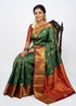 Indian woman draped in a bottle green semi Dharmavaram silk saree with a red zari border, seated on an antique wooden chair against a plain white studio backdrop.