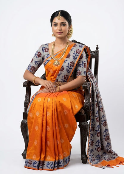 Traditional South Asian woman sitting on a wooden chair wearing a bright orange silk saree with a grey floral printed pallu and blouse