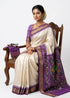 A traditional Indian woman sitting gracefully on a wooden chair, wearing an elegant cream silk saree with a vibrant lavender ikat-patterned pallu, gold zari border, and matching lavender blouse.