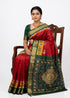 A beautiful woman seated on a carved wooden chair wearing a traditional deep crimson red silk saree with a dark green and gold zari border, featuring yellow accented bands, Patola-style elephant motifs, and a grand woven medallion pallu against a full white backdrop.