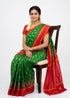 Traditional Indian woman wearing an emerald green and crimson red Pochampally Ikat pure silk saree seated on a wooden chair against a white background