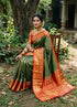 A graceful Indian woman seated in a lush traditional garden, wearing an elegant forest green and crimson red semi Dharmavaram silk saree with intricate gold zari borders and a rich brocade pallu.