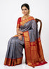 Traditional Indian woman seated on a chair wearing a slate gray Gadwal silk saree with a red zari pallu and red blouse against a plain white background.