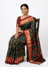 A beautiful woman sitting on a wooden chair, smiling and wearing a traditional bottle green Ikat silk saree with a contrasting maroon gold-zari border and matching blouse, styled with traditional Indian jewelry.