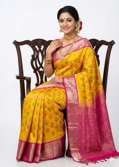 Model sitting on a carved wooden chair showcasing a vibrant yellow Kanchi Soft Silk saree with geometric arrow Ikat weave patterns and a contrasting traditional pink border and pallu with zari work against a white background.