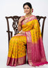 Model sitting on a carved wooden chair showcasing a vibrant yellow Kanchi Soft Silk saree with geometric arrow Ikat weave patterns and a contrasting traditional pink border and pallu with zari work against a white background.
