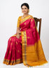 A traditional Indian woman sitting on a wooden chair wearing a Kanchi soft silk saree with a vibrant pink body featuring gold buta motifs, a rich yellow and gold zari pallu, and a matching yellow blouse, set against a solid white backdrop.