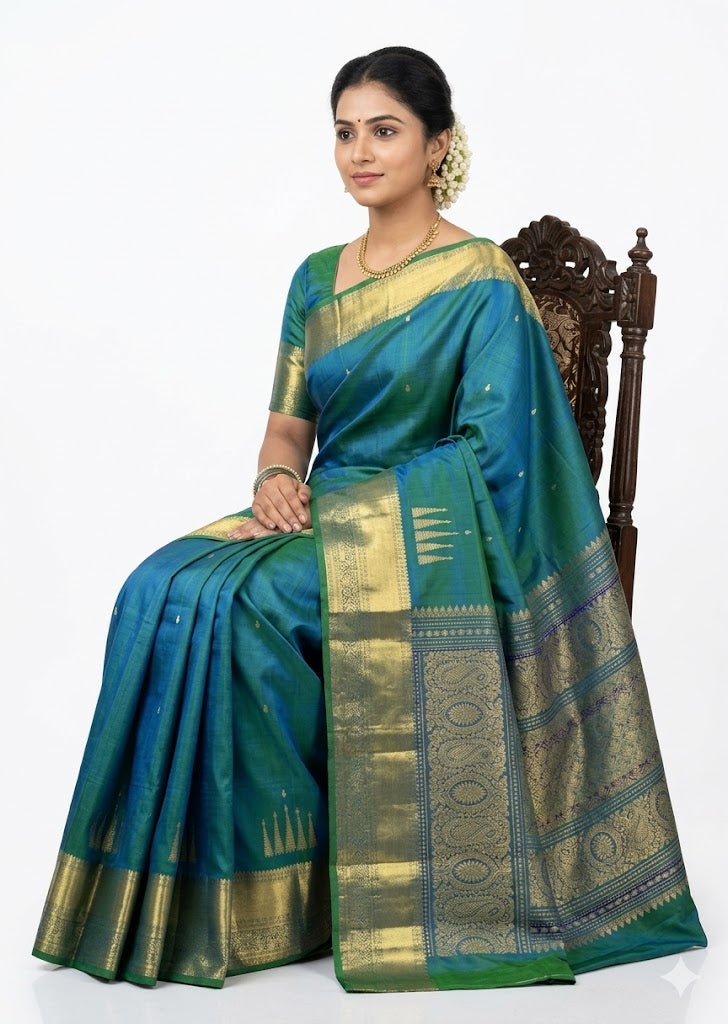 Traditional woman seated in an ornate chair, wearing a rich teal-blue Kanchipuram pattu saree with intricate gold zari work and adorned with gold jewelry. Find Kanchipuram Pattu Saree with Price here.