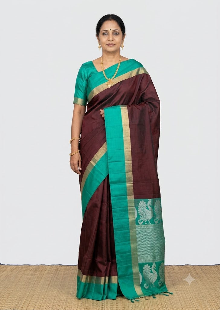 A beautiful Indian woman wearing a traditional deep maroon Kanchi Soft Silk Saree with a high-contrast teal green border featuring complex gold zari and silver peacock motifs.