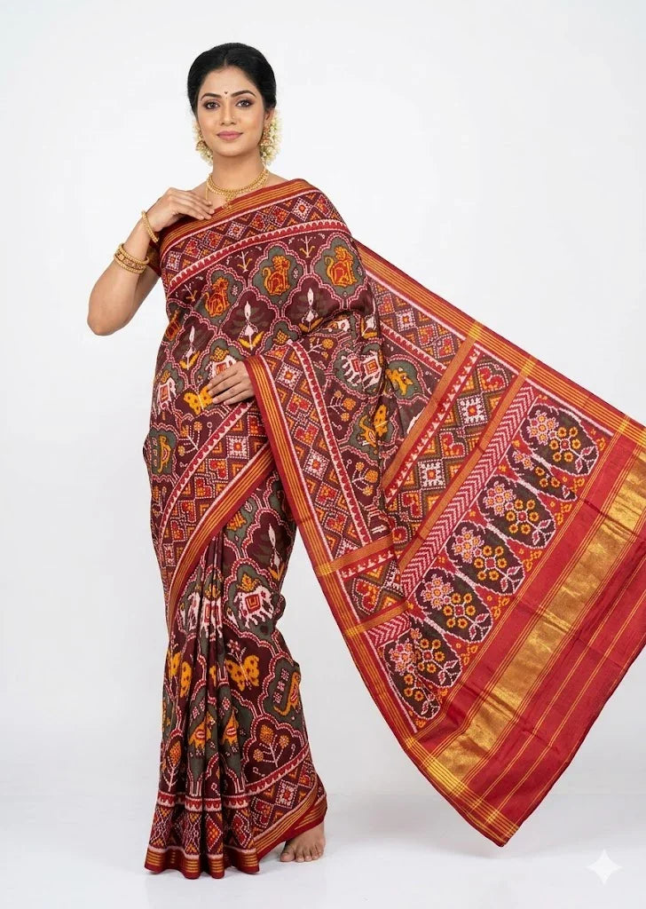A beautiful Indian woman standing elegantly against a white background, draped in a pure Patola silk saree. She is showcasing the deep maroon body with woven lion, elephant, and butterfly Ikat motifs, along with the rich red pallu featuring traditional floral and geometric patterns.