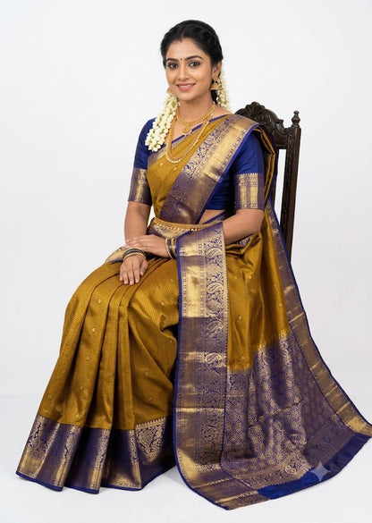 Indian woman wearing an antique mustard gold semi Dharmavaram silk saree with royal blue zari border, sitting gracefully on an antique wooden chair against a plain white studio background.