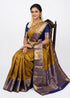 Indian woman wearing an antique mustard gold semi Dharmavaram silk saree with royal blue zari border, sitting gracefully on an antique wooden chair against a plain white studio background.