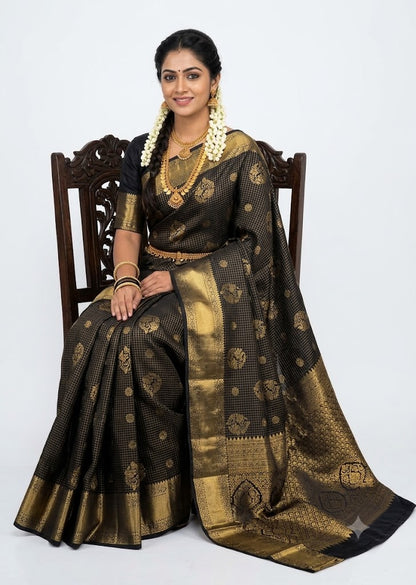 Indian woman wearing an antique mustard gold semi Dharmavaram silk saree with royal blue zari border, sitting gracefully on an antique wooden chair against a plain white studio background.