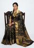 Indian woman wearing an antique mustard gold semi Dharmavaram silk saree with royal blue zari border, sitting gracefully on an antique wooden chair against a plain white studio background.