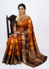 Indian woman wearing a mustard rust semi Dharmavaram silk saree with deep wine zari border, seated on an antique wooden chair against a plain white studio background