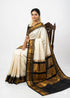 Traditional Indian woman sitting on a carved wooden chair, wearing an elegant off-white pure Gadwal silk saree featuring a contrasting black border and pallu with intricate gold zari temple motifs.