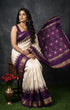 Indian woman seated wearing an elegant off-white ikkat silk saree with a royal purple gold zari border and temple design