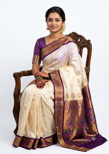 A beautiful smiling Indian woman seated on a carved wooden chair, wearing an elegant off-white Semi Gadwal Paithani saree with gold motifs and a purple border, perfectly paired with a contrasting dark lavender blouse.