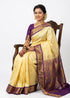 A smiling Indian woman seated gracefully on a carved wooden chair, wearing an elegant pale yellow Semi Gadwal Paithani saree with a rich purple and gold zari border, perfectly paired with a matching purple blouse against a white background.