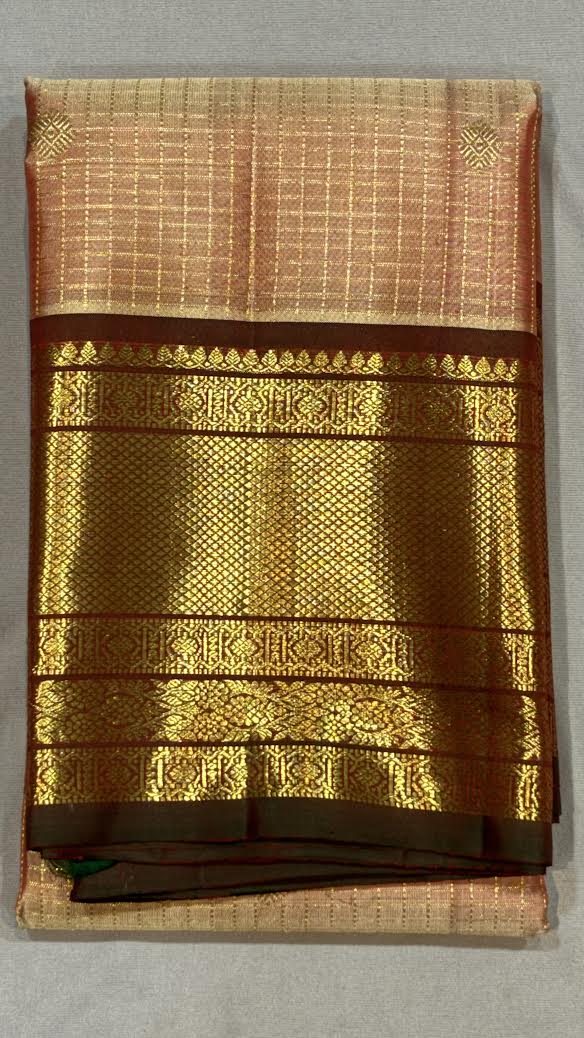A woman sitting on a wooden chair wearing a traditional Kanchipuram pure silk saree with a peach checkered body and a heavy dark maroon zari border.