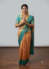Indian woman standing in a namaste pose wearing a traditional peach and teal blue silk saree with heavy gold zari border against a light grey studio backdrop.