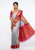 A traditional Indian model standing against a full white backdrop, wearing a premium silvery grey pure Gadwal silk saree with a vibrant deep pink and gold zari border,