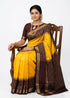 Beautiful Indian woman standing next to a wooden chair on a white backdrop, elegantly draped in a yellow traditional Pure Ikkat Silk saree with a chocolate brown border, pallu, and matching blouse.