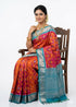 A beautiful Indian woman sitting on a classic wooden chair, showcasing a traditional orange Kanchi Soft Silk Ikat saree featuring a contrasting blue zari border on a clean white background.