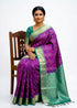 Model sitting on a wooden chair showcasing a traditional purple Kanchi Soft Silk Ikat saree with a contrasting Rama Green border and matching blouse on a solid white background.